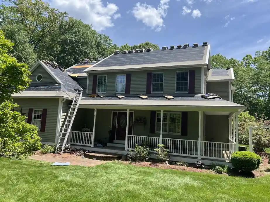 Colonial house mid re-roof with the old shingles stripped, felt paper down, ridge vents open and a ladder against the front porch